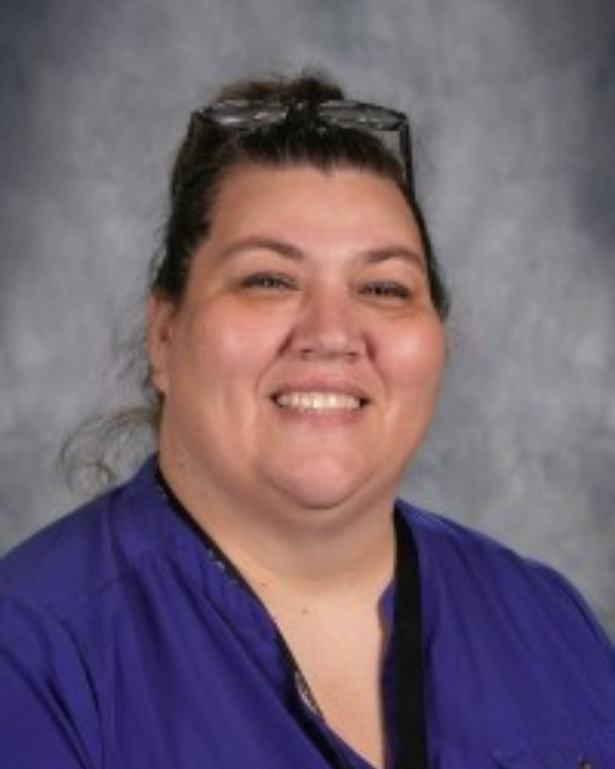 Smiling woman in a blue blouse against a gray studio backdrop, glasses resting on her head.