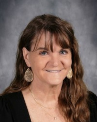 Portrait of a smiling woman with brown hair, gold earrings, and a black top against a gray studio backdrop.